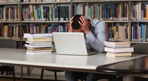 A man sitting at a library desk in front of a laptop and two stacks of books with his head in his hands and his face hidden by the laptop screen